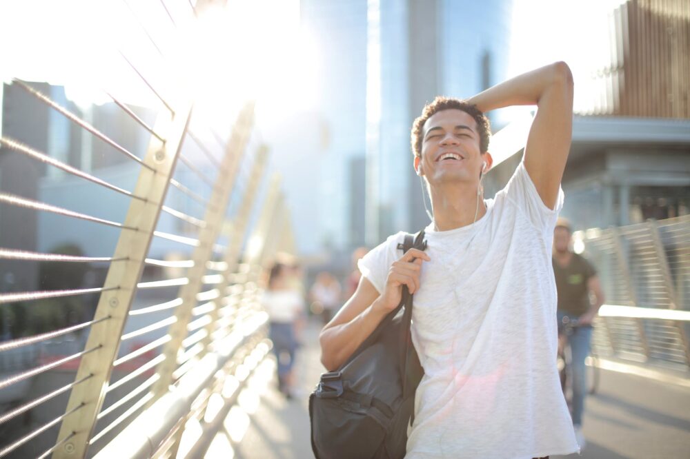 cheerful ethnic man in earphones with bag on shoulder in city in hot summer day